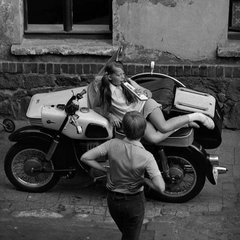 die Fotografie zeigt eine junge Frau, sich auf einem Motorrad reckelnd und einen Mann in Rückenansicht
